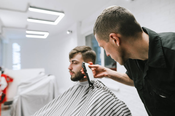 Parrucchiere maschile professionale taglia l'uomo barbuto nel moderno barbiere, utilizza clipper. Concentrarsi su un barbiere con un clipper crea un'acconciatura elegante per il cliente
. - Foto, immagini
