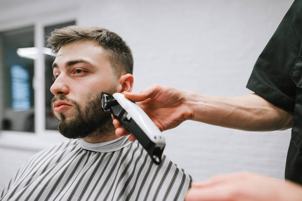 Barber trims the beard of a handsome man with a clipper. Closeup portrait of barber shop client. Barber correction of client's beard in men's hairdresser. - Fotoğraf, Görsel