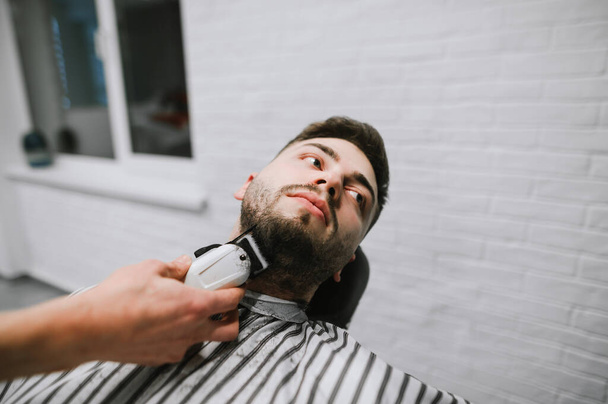 A borbély levágja egy jóképű férfi szakállát egy ollóval a kezében. Szoros portré a fodrászat ügyfeléről. Borbélykorrekció az ügyfél szakállán. Fodrász csinál frizura szakállas ügyfél borbély - Fotó, kép