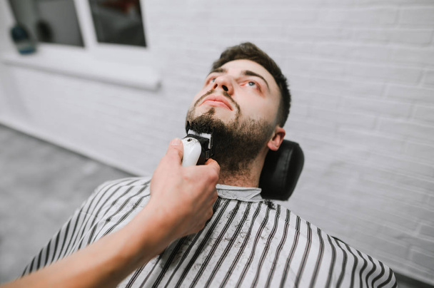 Professional barber with a hair clipper in his hand makes a beard hairstyle for the client. Man receives a service in a barber shop, hairdresser is engaged in the correction of his beard. - Foto, Bild