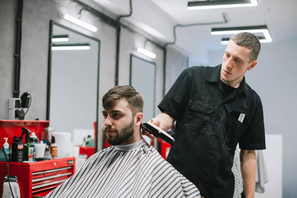 Barber trims client's hair with a clipper in his hands. Barber works in a barbershop workplace, makes a stylish hairstyle for a bearded man. Barber job concept. - Fotoğraf, Görsel