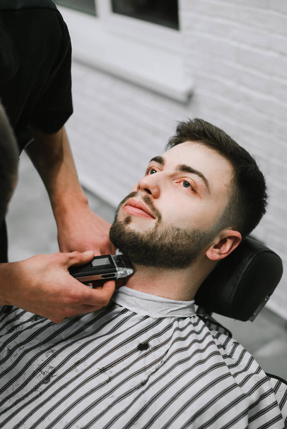Portrait of a man in barber shop, lying in a chair and looking up while the barber cuts his beard. Barber trims his beard seriously to the client. Vertical closeup portrait. - 写真・画像