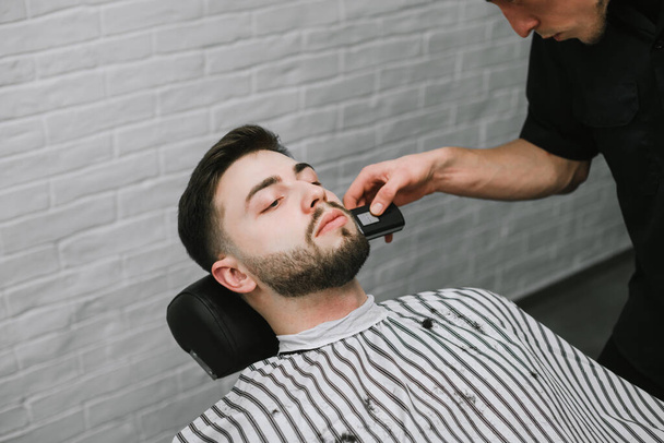 Professionele kapper maakt een baard trimmen een trimmer in zijn handen.Man met een baard is in een stoel met een serieus gezicht en gebruikt de kappers.Kapper snijdt de baard van de klant - Foto, afbeelding
