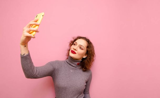 Cute lady z makijażem i kręcone rude włosy robi selfie na smartfonie na różowym tle, nosi szary sweter. Atrakcyjna nastolatka z czerwonymi ustami, pozująca na smartfonie, odizolowana. Przestrzeń kopiowania - Zdjęcie, obraz