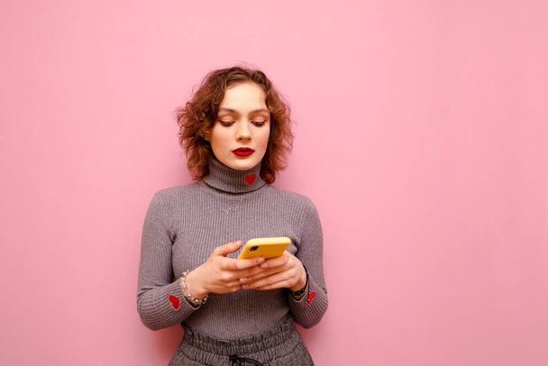Beautiful girl with curly hair wears a gray sweater, stands on a pink background and uses a smartphone. Cute teen girl writes a message on smartphone, isolated. Copy space - Photo, Image