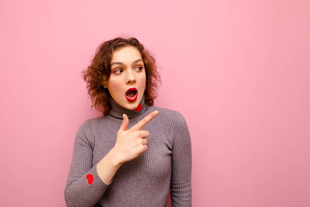 Surprised young girl in casual clothes and red curly hair shows pointing finger at copy space. Cute emotional lady showing thumbs up with surprised face on pink background. - 写真・画像