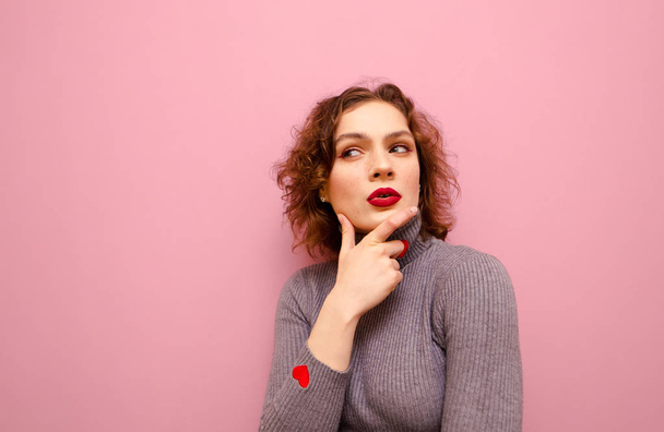 Funny curly girl with thoughtful face stands on pink background and looks away at Copy space and thinks. Closeup portrait of a thoughtful hipster girl in a gray sweater and with makeup. - 写真・画像