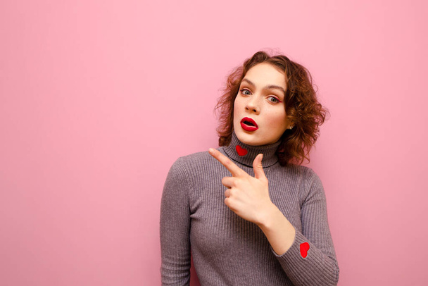 Portrait of surprised red-haired girl with curly hair on pink background, shows thumbs up at copy space and looks into camera with shocked face. Emotional teen girl shows empty space, looks surprised - Фото, зображення