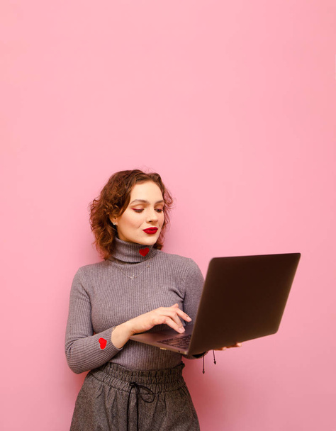 Vertical portrait of a curly red-haired girl in gray casual clothes uses a laptop on a pink background, looks into the screen with a serious face. Lady freelancer working on laptop ,isolated. - Fotoğraf, Görsel