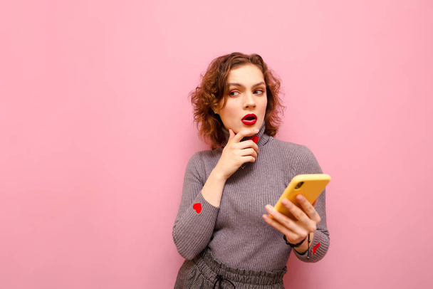 Pensive curly lady in gray clothes stands with smartphone in hand on pink background and looks away at copy space and thinks. Thoughtfully stylish girl with phone in hand, isolated. Copy space - Photo, Image