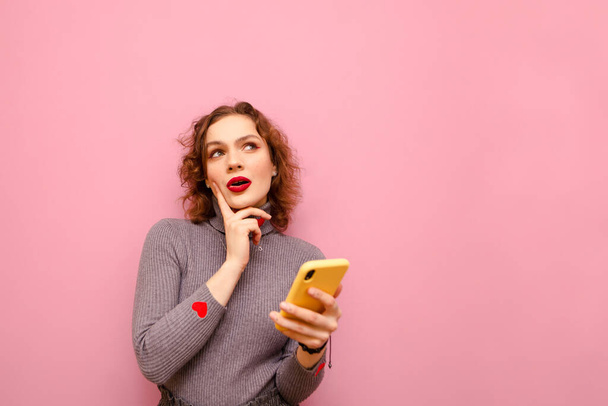 Pensive redhead curly girl in casual clothing and bright makeup stands with smartphone in hand on pink background and looks thoughtfully up at copy space. Isolated. - Photo, Image