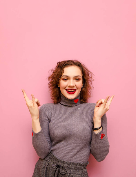 Smiling curly redhead girl isolated on pink background, shows heavy metal gesture and looks into camera with happy face, wears gray casual clothes with hearts. Vertical photo. - 写真・画像