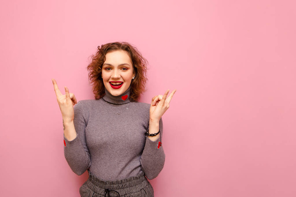 Happy cute girl with curly hair and casual clothes stands on a pink background and shows a heavy metal gesture, looks into the camera and smiles. Isolated. Rock music concept. - Fotoğraf, Görsel
