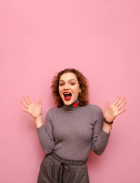 Studio portrait of joyful lady in casual clothes and with curly red hair on pastel pink background, looks into camera with surprised face and rejoices. Cheerful girl emotionally rejoices. Vertical - 写真・画像