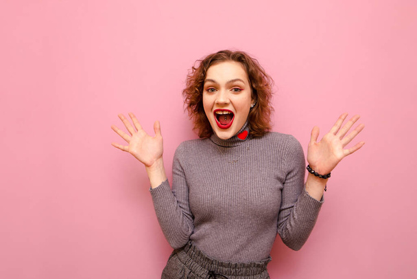 Portrait of joyful curly girl with red hair on pink pastel background, looks into camera with shocked face and raised hands. Happy girl rejoices, studio portrait. Isolated. - Фото, зображення