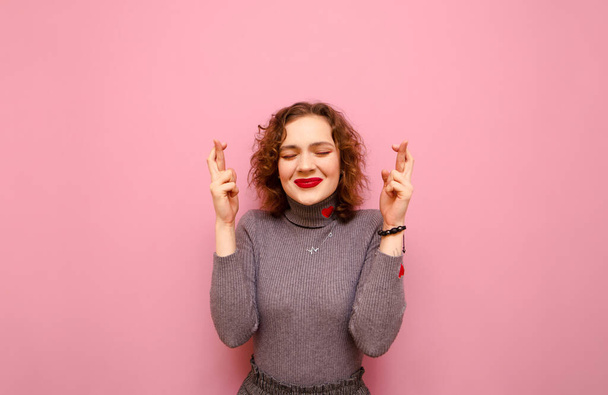 Cute girl with curly red hair crossed her fingers and closed her eyes against a pink background, smiling and making a wish.Funny lady with crossed fingers isolated on pink background with happy face - Foto, Bild