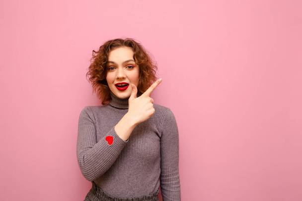 Positive girl in gray casual clothes and red curly hair stands on a pink background and points her finger to the side to advertise with a happy face. Isolated. Copy space - Foto, afbeelding