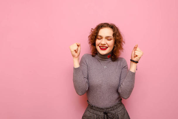 Positive teen girl with curly red hair and in a gray sweater stands with her eyes closed on a pink background and rejoices. Joyful young lady isolated on pink background. Copy space - Fotoğraf, Görsel