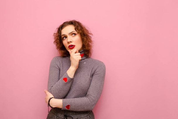 Attractive curly lady in casual clothes isolated on pastel pink background, looks away with pensive face on copy space. Pensive girl in sweater with hearts thinks on pink background. - Foto, Imagem