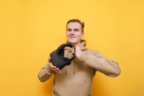 Cheerful young man isolated on yellow background with VR glasses in hands, looking into camera and smiling. The guy holds a virtual reality helmet and poses for the camera. VR concept. - Foto, Bild