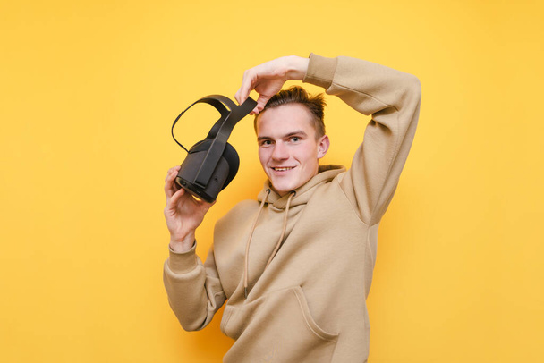 Portrait of happy guy on yellow background, standing with VR helmet in hands, looking into camera and smiling. Positive Young man trying on virtual reality helmet, isolated. VR gaming. - Φωτογραφία, εικόνα