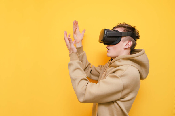 Portrait of surprised guy in VR helmet stands on yellow background and looks at his hands. Young man in casual clothes uses a virtual reality helmet. Isolated. - Photo, Image