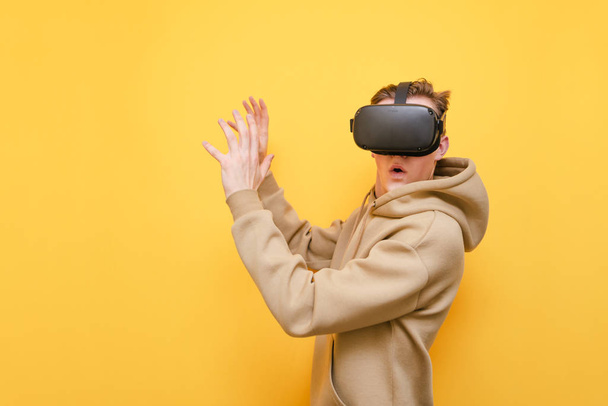 Surprised young man in VR clothes wearing VR glasses and with shocked face looks into camera on yellow background. Emotional guy is using Virtual Reality Helmet for the first time. Isolated. - Photo, Image