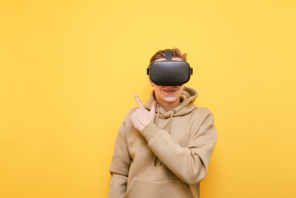Smiling young man in casual clothes wears VR glasses, stands on a yellow background and points his finger at Copy space. Happy guy in virtual reality helmet shows thumbs aside, isolated. - Photo, Image