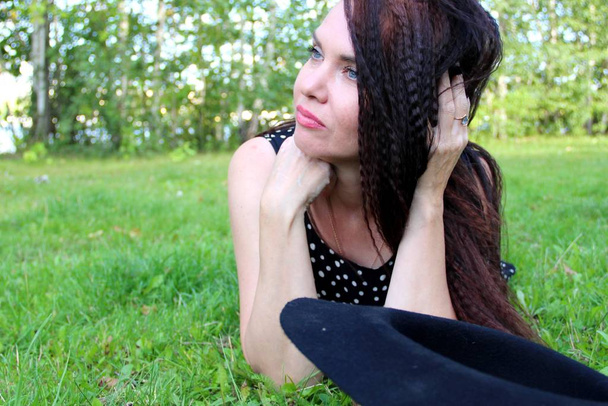 morena melancólica se encuentra en el césped verde en el parque. mira al lado alisando el pelo largo de la mano al lado del sombrero negro
 - Foto, Imagen