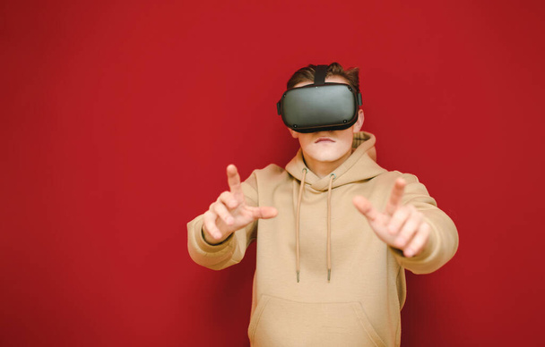 Vakava kaveri VR-kypärässä seisoo punaisella taustalla hymy kasvoillaan ja osoittaa kätensä kameraan. Nuori mies haluaa koskettaa kuvaa virtuaalitodellisuuden kypärässä
. - Valokuva, kuva