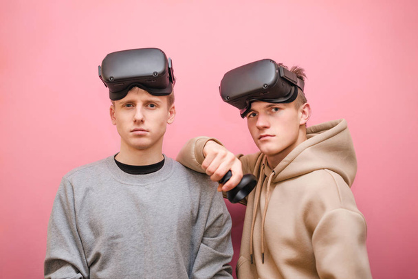Deux jeunes gars en réalité virtuelle casques isolés sur fond rose regardant dans la caméra avec un visage sérieux. Les joueurs adolescents dans les casques VR isolés sur fond rose
. - Photo, image