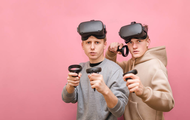 Portrait of two funny friends in VR helmet on a pink background looking in camera with a serious face. Two guys in casual clothes play VR games with controllers in their hands - 写真・画像