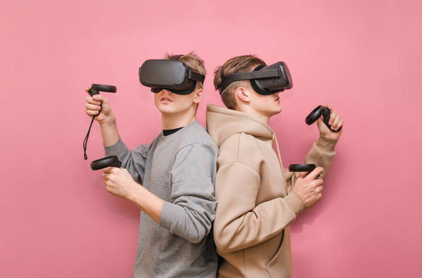 Zwei junge Männer mit Helmen auf dem Kopf stehen nebeneinander auf einem rosafarbenen Hintergrund, Controller in der Hand. Team von Jungs, die zusammen VR-Spiele spielen, isoliert. vr Gaming mit Freunden. - Foto, Bild