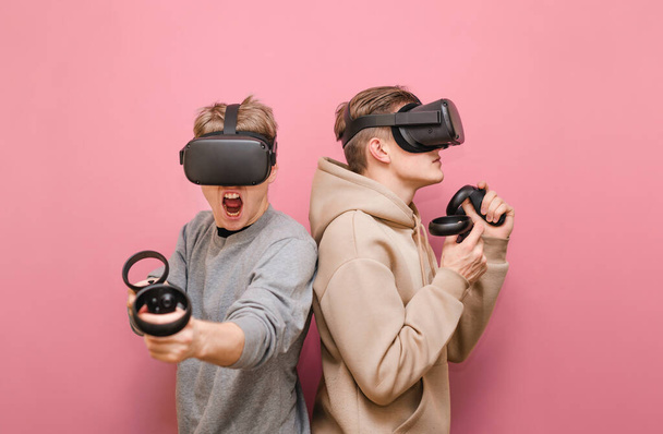 Couple d'amis drôles portant des casques VR et contrôleurs dans les mains, debout sur un fond rose et jouer à des jeux de réalité virtuelle. Deux jeunes hommes en vêtements décontractés jouant à des jeux en VR
 - Photo, image