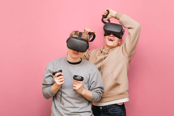 Two expressive friends in VR helmets compete in virtual reality games. Guy on the right is happy to win, another loser is upset by the defeat. Two guys playing VR games on a pink background. - Photo, Image