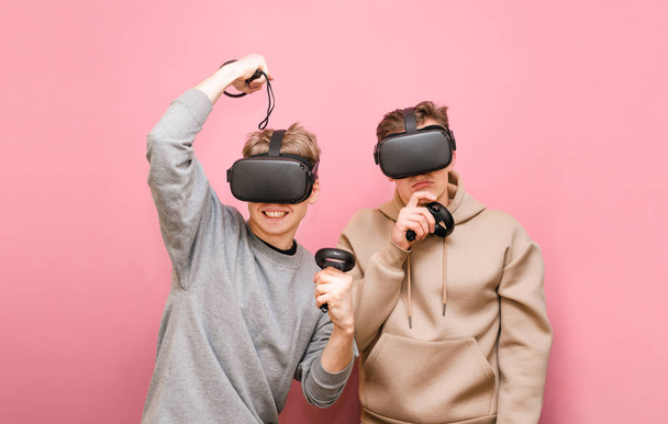 Two emotional friends spend time playing VR games, wearing virtual reality helmets and controllers in their hands and competing. Two guys in VR helmets isolated on pink background. - Photo, Image