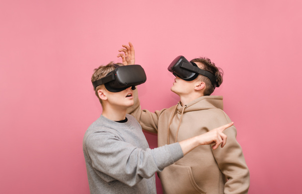 Two surprised guys in VR glasses stand on a pink background looking away and pointing their finger. Two others watching in headset of virtual reality, they are shocked. Copy space - Foto, Imagem