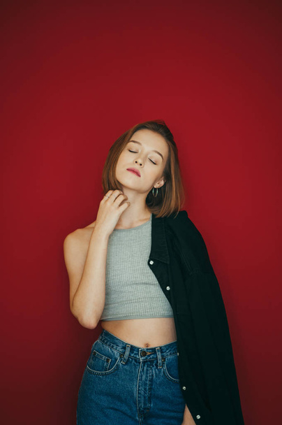 Portrait of sexy girl in t-shirt and shirt on red background, posing at camera with closed eyes. Attractive model poses on camera, closed her eyes with pleasure. Vertical photo. - Фото, зображення