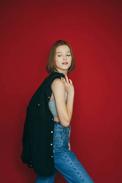 Vertical portrait of a fashionable girl in casual clothes stands on a red background with a jacket in her hands and poses at the camera. Stylish lady isolated on red background - Zdjęcie, obraz