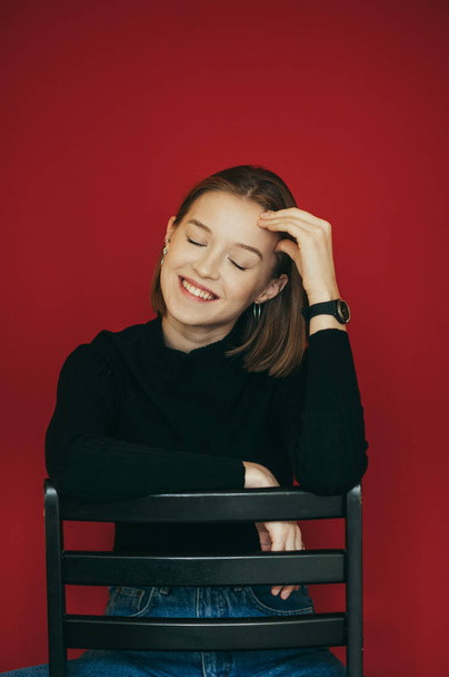 Happy girl model sits on a chair with her eyes closed in the studio on a red background and smiles sincerely.Joyful lady in black sweater sits on a chair against a red wall background. Vertical photo - Fotoğraf, Görsel