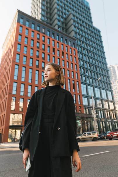 大都市の通りに流行の暗い服の中で魅力的な少女のライフスタイル写真、離れて見て。超高層ビルや通りを背景に街を歩くカジュアルな服装のファッショナブルな女性 - 写真・画像