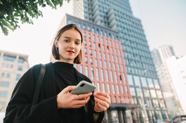 Portrait of a beautiful girl with a smartphone in her hands stands on the street of a metropolis against the background of a skyscraper building, looks into the camera and smiles. Copy space - Photo, Image