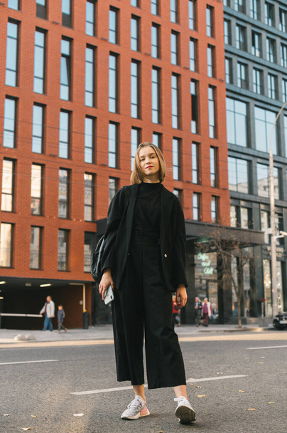 Portret van een meisje in stijlvolle casual kleding op een straat in de stad op zoek in camera met een serieus gezicht op de achtergrond van een modern gebouw.Jonge mode dame staat op de straat van een grote stad - Foto, afbeelding