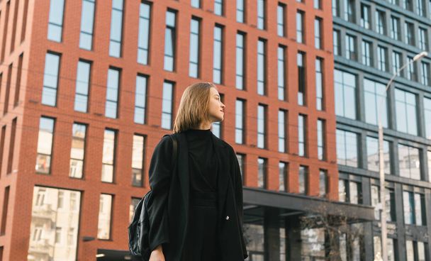 Fashionable girl in stylish casual clothes stands on the street against the backdrop of modern architecture and looks away. Street photo of fashionable lady in dark clothing. - Photo, Image