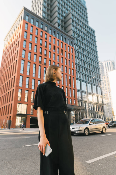 Vertical photo of a stylish lady in dark clothing stands with a smartphone against the backdrop of modern architecture and a metropolitan street, looking away. Lifestyle portrait of fashionable girl - Foto, Bild