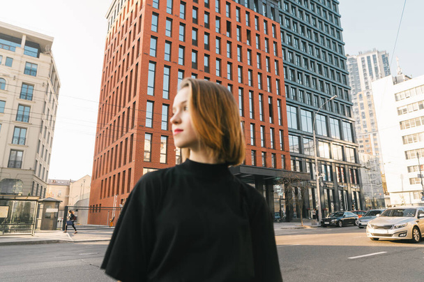 Blurred creative photo of attractive girl in dark clothes stands against the backdrop of a metropolis street and looks away. Focus on modern building. Abstract photo. Background - Foto, afbeelding
