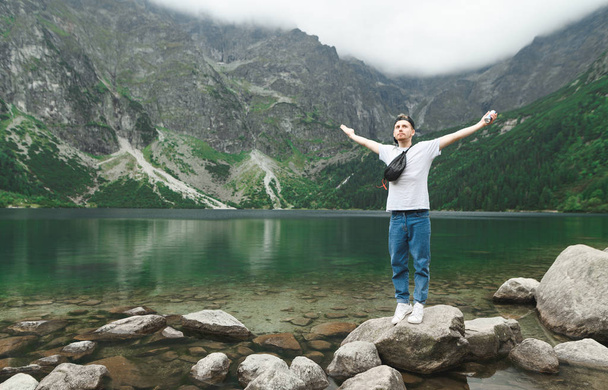 Young tourist in casual clothes stands on a rock on the shore of a mountain lake with his hands raised and enjoys the scenery.Young male tourist posing on the shores of Morskie Oko in Tatra Mountains - Photo, Image