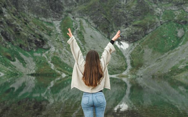 Радостная девушка в свитере стоит с поднятыми на фоне озера и горы руками, вид сзади. Хипстерская девушка, путешествующая в горах, позирует перед камерой с поднятыми руками. Женщина чувствует свободу в горах
 - Фото, изображение