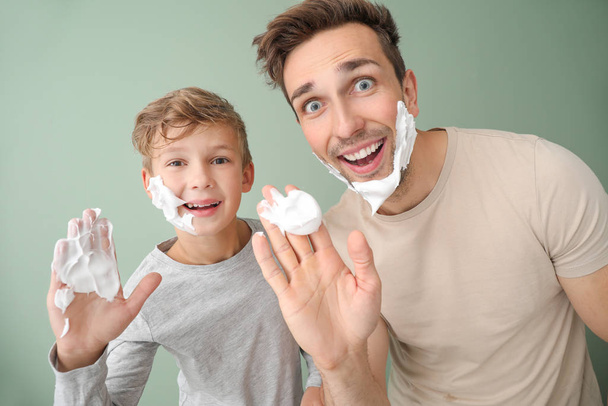 Funny father with son applying shaving foam onto their faces against color background - Photo, Image