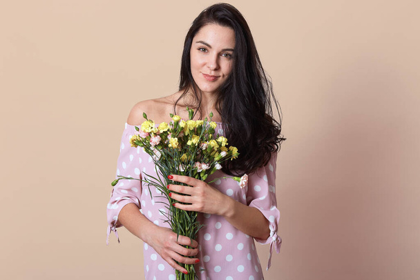 Studio portrait of miling Caucasian woman wearing pink dress, holding bouquet of flowers isolated over beige background, darkhaired female looking at camera. St. Valentines Day and present concept. - Фото, изображение